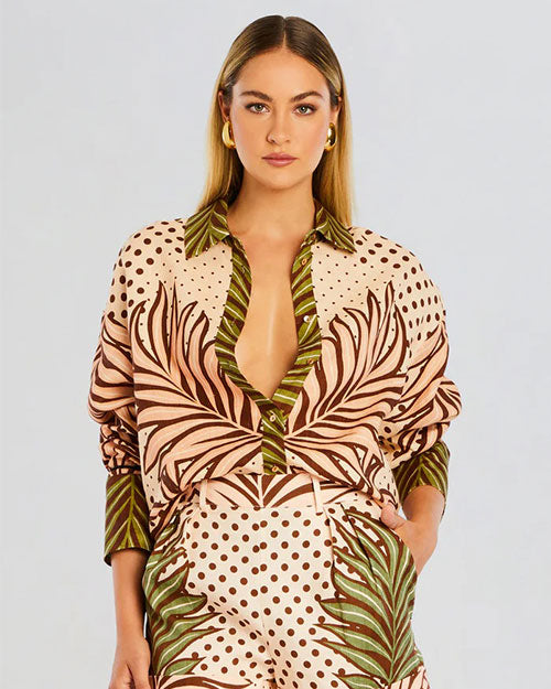 A knee-up view of a blonde model wearing a matching button-down shirt and bottoms with a polka-dot foliage pattern design, paired with blonde hoops. The background is plain white.