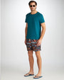 A full body shot of a model wearing a plain teal crew neck t-shirt with brown decorative shorts, square sunglasses, and blue flip flops. The model is standing in front of a white background.
