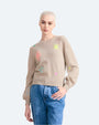 A knee-up view of a model with her thumb in her pocket, wearing a beige crewneck sweater with colorful abstract shapes scattered across it. The model is also wearing blue jeans and standing in front of a white background.