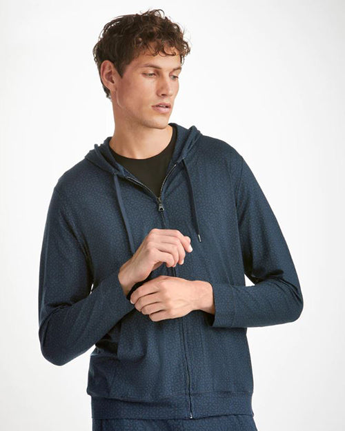 A waist-up view of a brunette model wearing a dark blue hooded zip-up jacket, featuring an all-over pattern of small, geometric shapes. The model is also wearing matching bottoms. The background is plain white.