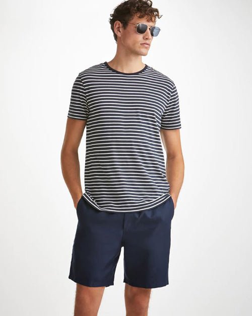 A knee-up view of a brunette model with his hands in his pockets wearing a white and black striped T-shirt with square sunglasses and navy shorts. The background is plain white.