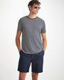 A knee-up view of a brunette model with his hands in his pockets wearing a white and black striped T-shirt with square sunglasses and navy shorts. The background is plain white.