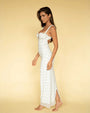 A sideview of a barefoot brunette model standing against a yellow background, wearing a long white form-fitting dress with thin straps and shoulder bows, featuring an empty space design at the chest, and a colorful pattern with a small blue scattered pattern.