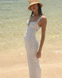 A brunette model on the beach wearing tan hat and a long white dress with thin straps and a small blue floral design with lacing at the bottom, and an empty space design at the chest.