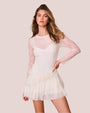 A knee-up view of a model with her hands on her hips, wearing a thin-strapped V-neck white dress with a sheer long-sleeve layer over it, and a ruffled skirt. The model is also wearing brown boots and standing in front of a white background.
