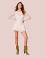 A full body shot of a model with her hands on her hips, wearing a thin-strapped V-neck white dress with a sheer long-sleeve layer over it, and a ruffled skirt. The model is also wearing brown boots and standing in front of a white background.