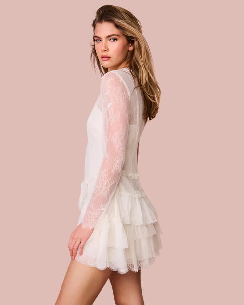 A knee-up sideview of a blonde model wearing a thin-strapped V-neck white dress with a sheer long-sleeve layer over it, and a ruffled skirt. The background is plain white.