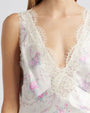 A close-up chest-level view of a model wearing a white floral-patterned tank top adorned with pink flowers, a V-neck, and delicate lace trim with scalloped edges along the neckline and straps.