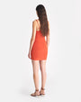The backside view of a person wearing an orangish/red sleeveless bodycon dress that falls above the knees. The dress has thin straps and a square neckline, and the person is also wearing strappy high-heeled sandals. The background is plain white.