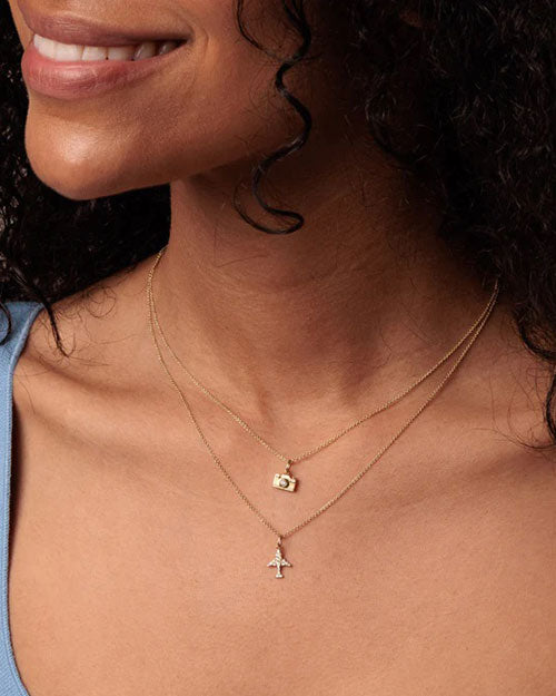 A close-up image of a smiling model's upper chest and chin area wearing two gold necklaces: a shorter one with a small camera charm and a longer one with an airplane charm adorned in jewels.