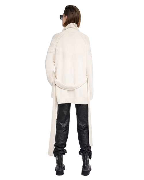 Person standing with their back to the camera, wearing a white oversized high-collar sweater, black textured pants, black boots, and sunglasses; short hair visible.