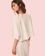 The backside view of a brunette model wearing a slightly cropped, white, loose-fitting top and a matching white skirt. The background is plain and pink.