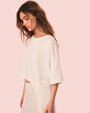 A side view of a brunette model wearing a cropped, white, loose-fitting top and a matching white skirt. The background is plain and pink.