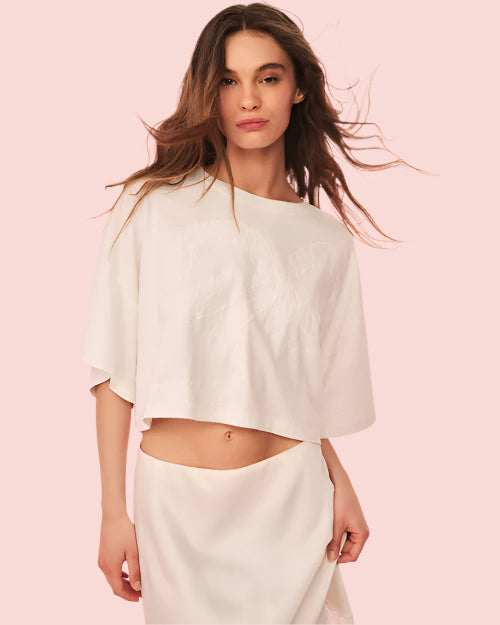 A brunette model with her hair blowing behind her, wearing a cropped, white loose-fitting top and a matching white skirt. The background is plain and pink.
