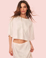 A brunette model with her hair blowing behind her, wearing a cropped, white loose-fitting top and a matching white skirt. The background is plain and pink.