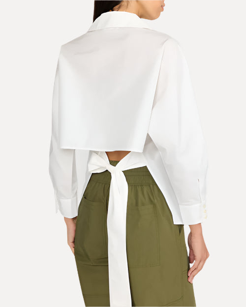 A model wearing a white long-sleeve top that features an open back design at the lower back, tied together with a long delicate bow. She is wearing army green pants to match, and standing against a plain white background.