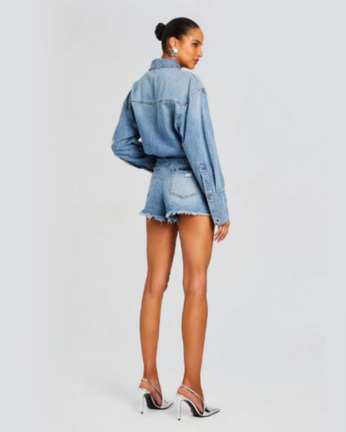 A full body shot of a brunette model wearing a bun, and a denim romper designed as a collared shirt tucked into blue denim shorts with frayed hems. The model is also wearing pointed toe silver heels and standing in front of a plain white. The background is plain white.