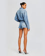 A full body shot of a brunette model wearing a bun, and a denim romper designed as a collared shirt tucked into blue denim shorts with frayed hems. The model is also wearing pointed toe silver heels and standing in front of a plain white. The background is plain white.