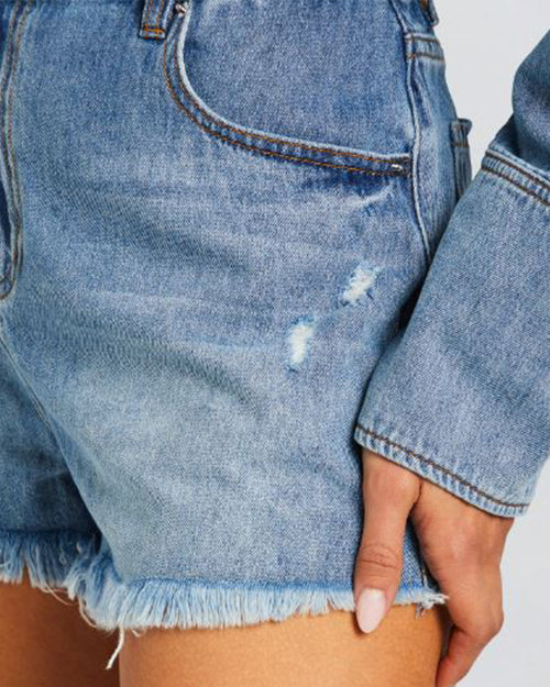 A close-up hip-level view of a model with nude painted nails and her hand at her hips, wearing a denim shirt with long cuffs, and denim shorts with frayed hems.