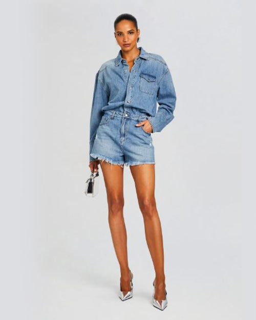 A full body shot of a brunette model wearing a denim romper designed as a button-down shirt tucked into blue denim shorts with frayed hems. The model is also holding a silver purse and wearing pointed toe silver heels. The background is plain white.