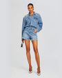 A full body shot of a brunette model wearing a denim romper designed as a button-down shirt tucked into blue denim shorts with frayed hems. The model is also holding a silver purse and wearing pointed toe silver heels. The background is plain white.