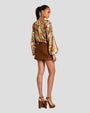 The backside view of a person standing against a plain background wearing a long-sleeved, floral patterned blouse with earthy tones, a deep neckline and a short brown suede skirt. The model is standing in front of a white background, accessorizing with brown high-heels and gold jewelry.