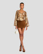 A complete frontal image of a person standing against a plain background wearing a long-sleeved, floral patterned blouse with earthy tones, and a deep neckline with a cut-out design and a bow at the and a short brown suede skirt. The person is also seen wearing brown heels, a small white bag and gold hoop earrings.