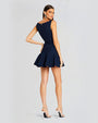 An angled full body view of a brunette model wearing a textured blue sleeveless dress with a short lightly ruffled skirt. She is accessorizing with large silver earrings and silver heels. The model is standing in front of a white background.
