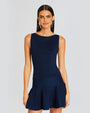 A knee-up view of a brunette model wearing a textured blue sleeveless dress with a short lightly ruffled skirt. She is accessorizing with large silver earrings and is standing in front of a white background.