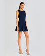 An angled full body view of a brunette model wearing a textured blue sleeveless dress with a short lightly ruffled skirt. She is accessorizing with large silver earrings and silver heels. The model is standing in front of a white background.