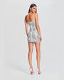 A full body backside view of a brunette model with her hair swept back wearing a short, grey thin strapped dress adorned with shimmering sequins. The model is also wearing dangly silver earrings with white heels and is standing in front of a white background.