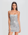 A knee-up view of a brunette model wearing a short, grey thin strapped dress adorned with shimmering sequins. The model is also wearing long dangly silver earrings and is standing in front of a white background.