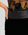 A close-up hip-level view of a person wearing a black dress with intricate floral embroidery on the sheer bodice. The person's left hand is resting on their hip, and the dress has a wide, black waistband that cinches at the waist.