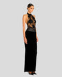 An angled full body view of a brunette model with a bun wearing a long, black dress with a sheer, floral-patterned bodice and halter neckline, with a solid black skirt that extends to the floor. The model is also wearing black high-heeled shoes and is standing in front of a white background.