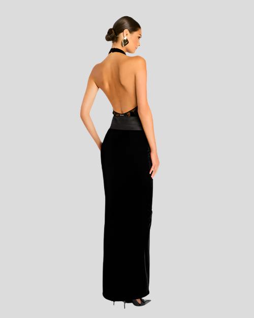 An angled backside view of a brunette model with a bun wearing a long black dress with an open-back and a halter neckline, with a solid black skirt that extends to the floor. The model is also wearing black high-heeled shoes and large diamond shaped silver earrings. The model is standing in front of a white background.