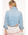 A waist-up backside view of a blonde model wearing a light-blue denim jacket with the collar popped, and a brown square patch at the bottom corner. The model is wearing a white lace embroidered dress with frills and red cherry designs and stripes around the edges. The background is plain white.