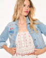 A zoomed in waist-up view of a blonde model holding open a light-blue denim jacket with cherries and a bow embroidered above the right chest pocket. The model is wearing a white lace embroidered top with frills and cherry designs around the edges. The background is plain white.
