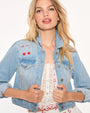 A waist-up view of a blonde model holding onto the front of a light-blue denim jacket with cherries and a bow embroidered above the right chest pocket. The model is wearing a white lace embroidered top with frills and cherry designs around the edges. The background is plain white.