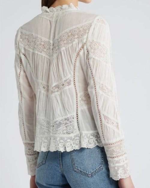 A close-up backside view of a model wearing a white, long-sleeved lace blouse with intricate lace designs and scalloped edges at the hem and cuff. She is wearing light blue jeans and standing in front of a white background.