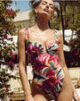 An upper body shot of a model by at the poolside, surrounded by foliage and beige chairs, wearing a vibrant one-piece swimsuit with a colorful watercolor floral pattern, with a sweetheart neckline and a small cutout detail.