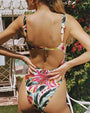 A backside, upper body shot of a brunette model with her hands on her hips standing by the poolside, surrounded by foliage and beige chairs, wearing a vibrant one-piece swimsuit with a colorful watercolor floral pattern, with a sweetheart neckline and a small cutout detail.