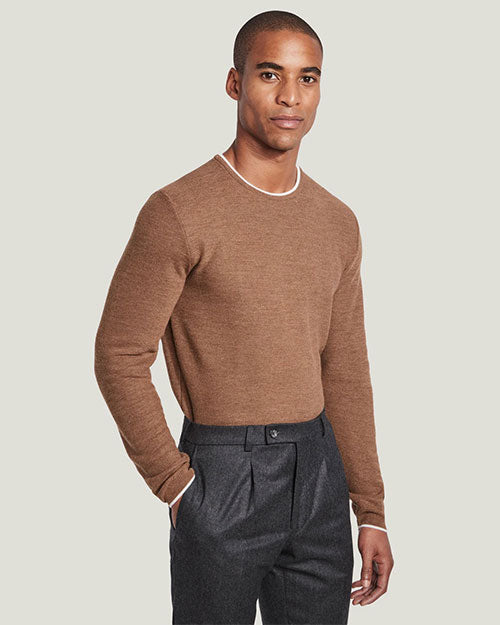 A knee-up view of a model with his hand in his pocket wearing a light-brown crewneck sweater with a white trim tucked into a pair of dark grey trousers. The background is plain white.