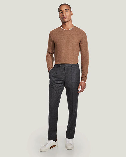 A full body view of a model with his hand in his pocket wearing a light-brown crewneck sweater with a white trim tucked into a pair of dark grey trousers with white sneakers. The background is plain white.
