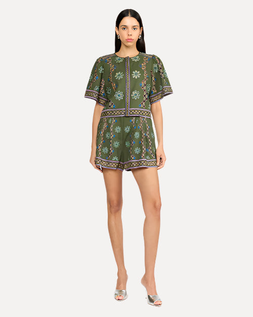 A model is wearing a green short-sleeved top with matching green shorts that feature a vibrant floral pattern. The pattern features vertical stripe designs and horizontal bands around the leg openings and a wide belt made of the same fabric, tied in a large bow at the waist. The person is also wearing silver open-toe high-heeled shoes, standing in front of a white background.