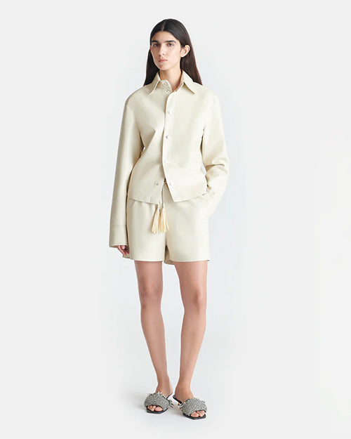 A brunette model wearing an Off-White long-sleeved button-down jacket with matching shorts and tasseled drawstrings. The person is also wearing slip-on shoes with textured details, standing in front of a plain background.