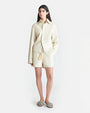 A brunette model wearing an Off-White long-sleeved button-down jacket with matching shorts and tasseled drawstrings. The person is also wearing slip-on shoes with textured details, standing in front of a plain background.