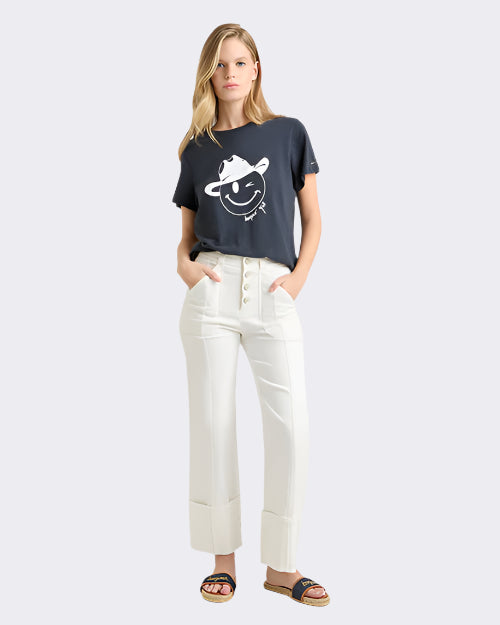 A blonde model standing against a white background with her hands in her pockets, wearing a navy t-shirt with a graphic of a smiling winky face wearing a cowboy hat. The model is also wearing white high-waisted pants with multiple buttons down the fly cuffs at the bottom, along with navy open-toed sandals. The background is plain white.