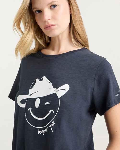 A close-up torso-up shot of a blonde model wearing a navy crewneck t-shirt with a graphic of a smiling winky face wearing a cowboy hat. The shirt has the words "bonjour y'all" written in cursive below the face.