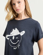 A close-up torso-up shot of a blonde model wearing a navy crewneck t-shirt with a graphic of a smiling winky face wearing a cowboy hat. The shirt has the words "bonjour y'all" written in cursive below the face.