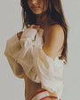 A torso only shot of a model wearing and holding up to her chest a white shirt hanging off one shoulder, and light red stitching. The model is also wearing white underwear with red lining and standing against a grey background.